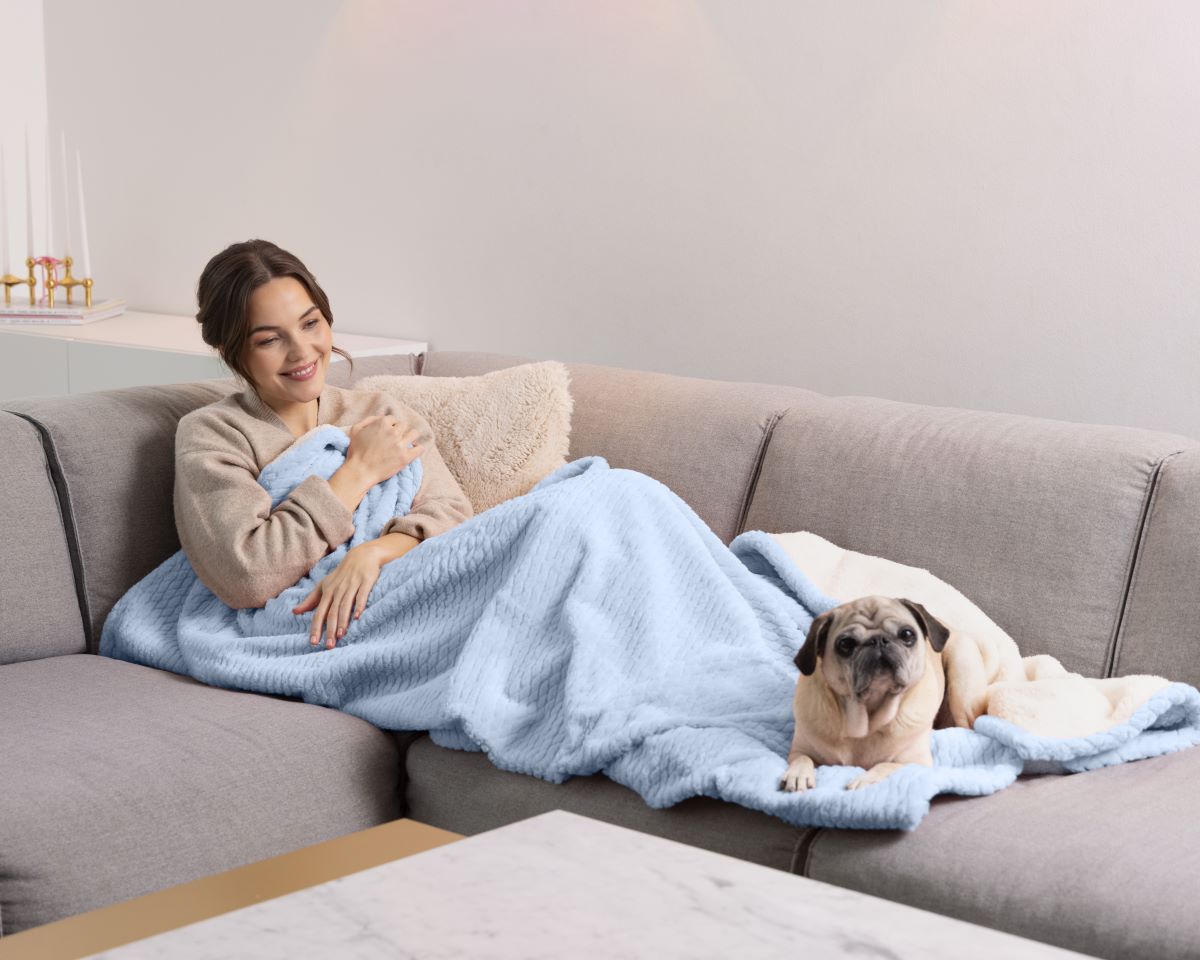 Woman relaxing on the sofa with a light blue medisana heated blanket, with a dog lying beside her.